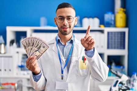 Téléchargez les photos : Young hispanic man working at scientist laboratory holding money surprised with an idea or question pointing finger with happy face, number one - en image libre de droit
