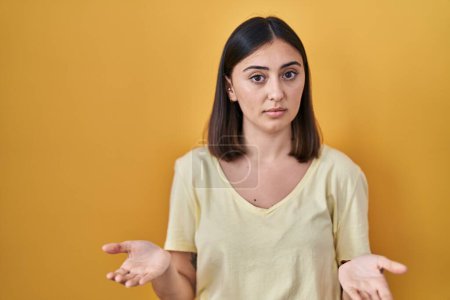 Foto de Hispanic girl wearing casual t shirt over yellow background clueless and confused with open arms, no idea concept. - Imagen libre de derechos