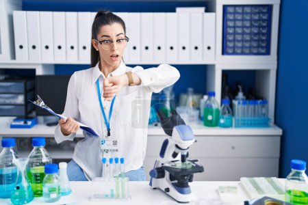 Téléchargez les photos : Jeune femme brune travaillant au laboratoire scientifique regardant le temps de la montre inquiet, peur de se retrouver en retard - en image libre de droit