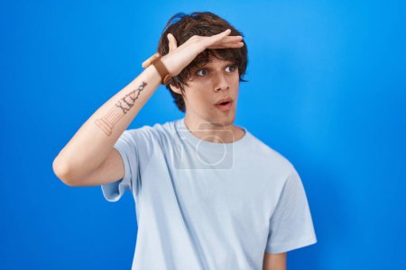 Photo for Hispanic young man standing over blue background very happy and smiling looking far away with hand over head. searching concept. - Royalty Free Image