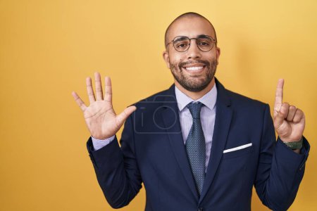 Foto de Hombre hispano con barba vistiendo traje y corbata mostrando y señalando con los dedos número seis mientras sonríe confiado y feliz. - Imagen libre de derechos