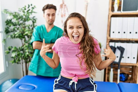 Photo for Young hispanic woman at physiotherapist appointment shouting with crazy expression doing rock symbol with hands up. music star. heavy concept. - Royalty Free Image