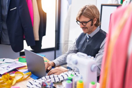 Téléchargez les photos : Young blond man tailor smiling confident using laptop at clothing factory - en image libre de droit
