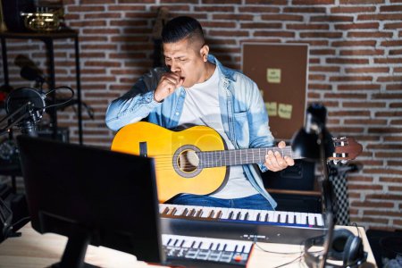Téléchargez les photos : Jeune homme hispanique jouant de la guitare classique au studio de musique se sentant mal et toussant comme symptôme de rhume ou de bronchite. concept de soins de santé. - en image libre de droit