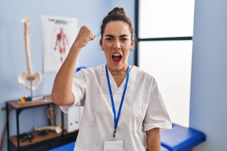 Photo for Young hispanic woman working at rehabilitation clinic angry and mad raising fist frustrated and furious while shouting with anger. rage and aggressive concept. - Royalty Free Image