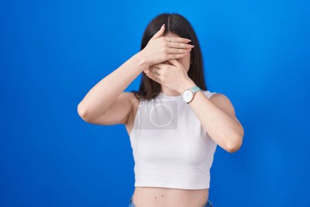 Foto de Young caucasian woman standing over blue background covering eyes and mouth with hands, surprised and shocked. hiding emotion - Imagen libre de derechos