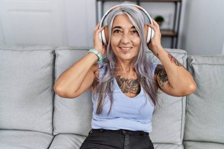 Foto de Mujer de pelo gris de mediana edad sonriendo confiada escuchando música en casa - Imagen libre de derechos