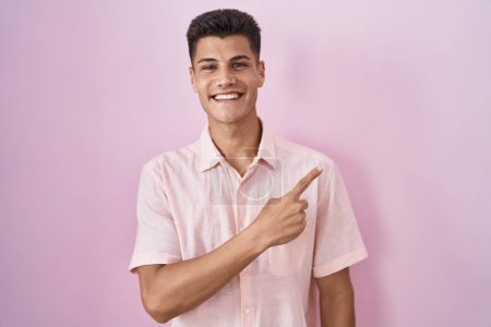 Photo for Young hispanic man standing over pink background cheerful with a smile on face pointing with hand and finger up to the side with happy and natural expression - Royalty Free Image