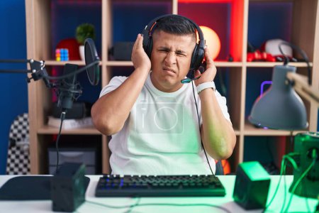Foto de Joven hispano jugando videojuegos cubriendo oídos con dedos con expresión molesta por el ruido de la música alta. concepto de sordo. - Imagen libre de derechos