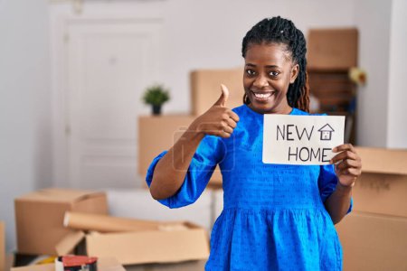Foto de Mujer africana con el pelo trenzado moviéndose a una nueva casa sosteniendo pancarta sonriendo feliz y positivo, pulgar hacia arriba haciendo excelente y signo de aprobación - Imagen libre de derechos