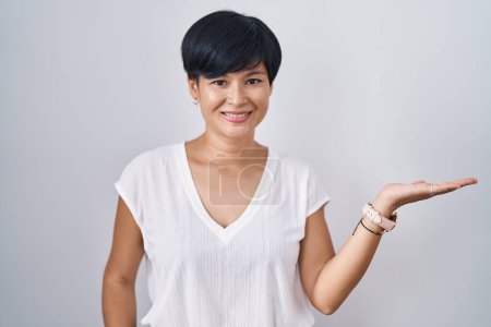 Téléchargez les photos : Jeune femme asiatique avec les cheveux courts debout sur fond isolé souriant gai présentation et pointant avec la paume de la main en regardant la caméra. - en image libre de droit