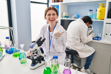 Foto de Joven dos personas que trabajan en el laboratorio científico señalando el pulgar hacia el lado sonriendo feliz con la boca abierta - Imagen libre de derechos