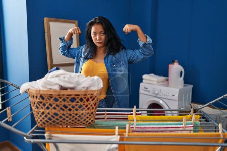 Foto de Young asian woman hanging clothes at clothesline showing arms muscles smiling proud. fitness concept. - Imagen libre de derechos