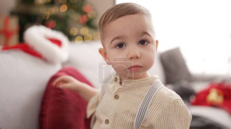Foto de Adorable niño de pie junto al árbol de Navidad con expresión seria en casa - Imagen libre de derechos