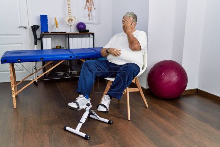 Photo for Senior caucasian man at physiotherapy clinic using pedal exerciser with sad expression covering face with hands while crying. depression concept. - Royalty Free Image