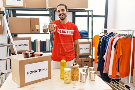 Photo for Young hispanic man wearing volunteer t shirt at donations stand pointing to you and the camera with fingers, smiling positive and cheerful - Royalty Free Image