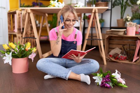 Foto de Joven florista rubia hablando en el libro de lectura de teléfonos inteligentes en la tienda de flores - Imagen libre de derechos