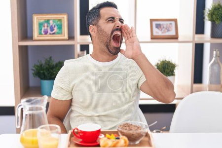 Photo for Hispanic man with beard eating breakfast clueless and confused with open arms, no idea and doubtful face. - Royalty Free Image