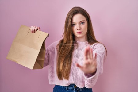 Foto de Young caucasian woman holding shopping bag and credit card with open hand doing stop sign with serious and confident expression, defense gesture - Imagen libre de derechos