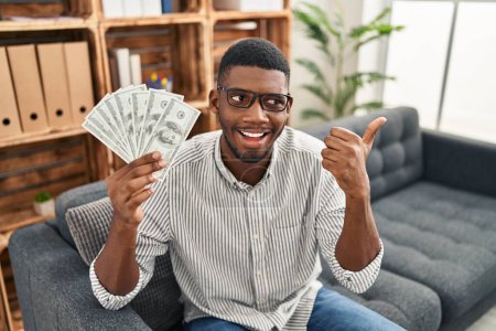 Photo for African american man holding dollars pointing thumb up to the side smiling happy with open mouth - Royalty Free Image