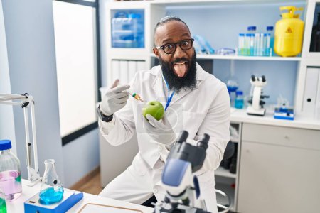 Photo for African american man working at scientist laboratory with apple sticking tongue out happy with funny expression. - Royalty Free Image