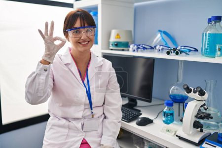 Foto de Mujer morena joven que trabaja en el laboratorio científico sonriendo positiva haciendo signo de ok con la mano y los dedos. expresión exitosa. - Imagen libre de derechos