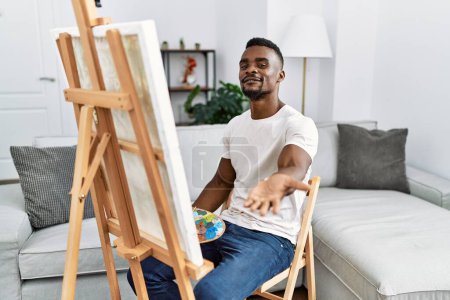 Téléchargez les photos : Jeune homme africain peignant sur toile à la maison souriant joyeuse offrant main de paume donnant assistance et acceptation. - en image libre de droit