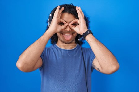 Foto de Hombre hispano con el pelo rizado escuchando música usando auriculares haciendo un buen gesto como prismáticos sacando la lengua, ojos mirando a través de los dedos. expresión loca. - Imagen libre de derechos