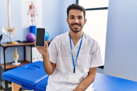 Foto de Young hispanic physiotherapist man holding smartphone at the clinic looking positive and happy standing and smiling with a confident smile showing teeth - Imagen libre de derechos
