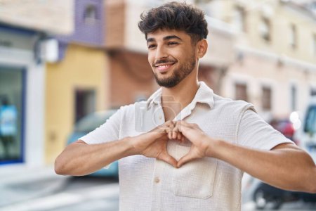 Téléchargez les photos : Jeune homme arabe souriant confiant faire geste de coeur avec les mains à la rue - en image libre de droit