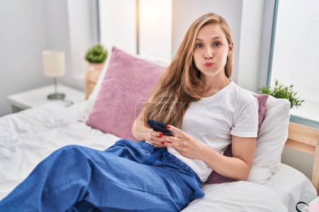 Foto de Young blonde woman using smartphone on bed puffing cheeks with funny face. mouth inflated with air, catching air. - Imagen libre de derechos