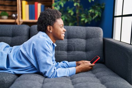 Téléchargez les photos : Femme afro-américaine utilisant un smartphone couché sur le canapé à la maison - en image libre de droit