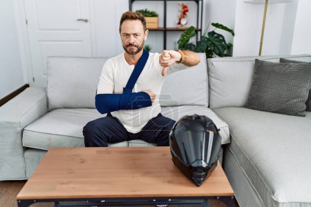 Téléchargez les photos : Middle age man with beard wearing arm on sling because motorbike accident with angry face, negative sign showing dislike with thumbs down, rejection concept - en image libre de droit
