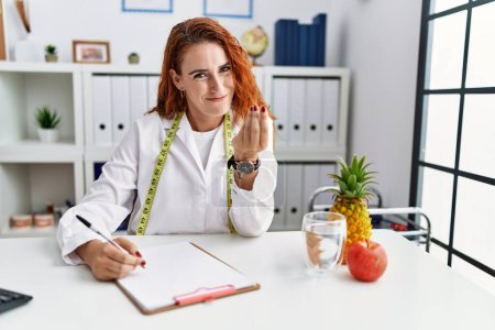 Photo for Young redhead woman nutritionist doctor at the clinic doing money gesture with hands, asking for salary payment, millionaire business - Royalty Free Image