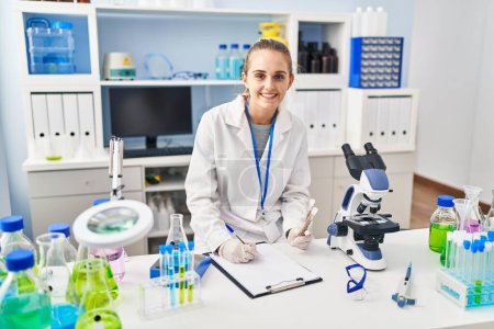 Foto de Young blonde woman wearing scientist uniform holding test tubes writing on checklist at laboratory - Imagen libre de derechos