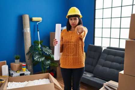 Photo for Young hispanic woman standing on new home with blueprints pointing with finger to the camera and to you, confident gesture looking serious - Royalty Free Image