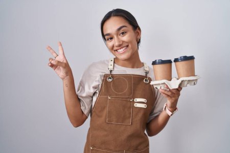 Photo for Young hispanic woman wearing professional waitress apron holding coffee smiling looking to the camera showing fingers doing victory sign. number two. - Royalty Free Image