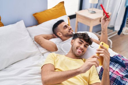 Téléchargez les photos : Couple de deux hommes utilisant un smartphone couché sur le lit dans la chambre - en image libre de droit