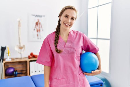 Téléchargez les photos : Young caucasian woman wearing physiotherapist uniform holding fit ball at physiotherapy clinic - en image libre de droit