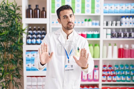 Téléchargez les photos : Handsome hispanic man working at pharmacy drugstore moving away hands palms showing refusal and denial with afraid and disgusting expression. stop and forbidden. - en image libre de droit
