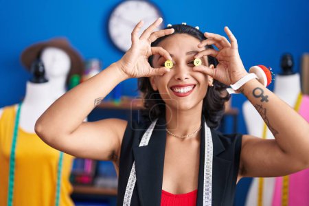 Téléchargez les photos : Young beautiful hispanic woman tailor smiling confident holding buttons over eyes at clothing factory - en image libre de droit