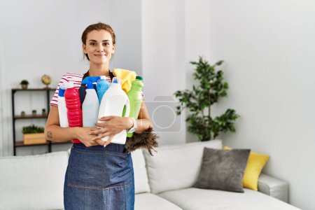 Foto de Mujer joven sonriendo confiada sosteniendo productos de limpieza en casa - Imagen libre de derechos