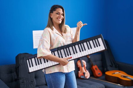 Téléchargez les photos : Blonde woman holding piano keyboard pointing thumb up to the side smiling happy with open mouth - en image libre de droit