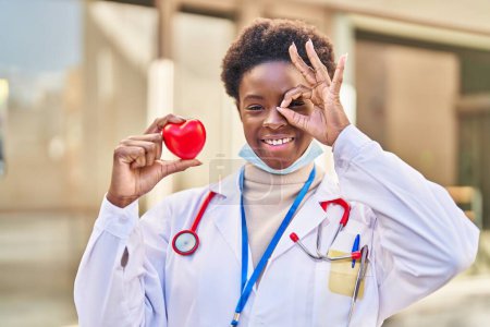 Foto de Mujer afroamericana vistiendo uniforme médico sosteniendo el corazón sonriendo feliz haciendo signo de bien con la mano en el ojo mirando a través de los dedos - Imagen libre de derechos