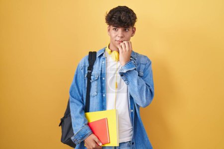 Téléchargez les photos : Adolescent hispanique portant un sac à dos étudiant et tenant des livres à l'air stressé et nerveux avec les mains sur les ongles mordants de la bouche. problème d'anxiété. - en image libre de droit