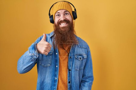 Téléchargez les photos : Homme caucasien à longue barbe écoutant de la musique à l'aide d'écouteurs faisant des pouces heureux geste avec la main. approuvant l'expression regardant la caméra montrant le succès. - en image libre de droit
