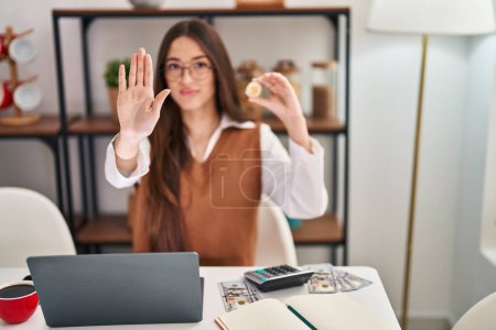 Téléchargez les photos : Jeune femme brune travaillant avec ordinateur portable tenant monnaie virtuelle Bitcoin avec main ouverte faisant stop signe avec expression sérieuse et confiante, geste de défense - en image libre de droit