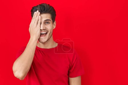 Photo for Young hispanic man standing over red background covering one eye with hand, confident smile on face and surprise emotion. - Royalty Free Image