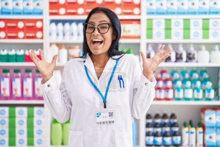 Téléchargez les photos : Hispanic woman working at pharmacy drugstore celebrating victory with happy smile and winner expression with raised hands - en image libre de droit
