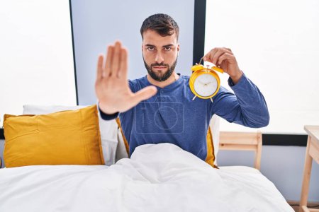 Photo for Handsome hispanic man in the bed holding alarm clock with open hand doing stop sign with serious and confident expression, defense gesture - Royalty Free Image